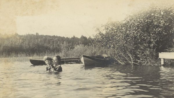 Eleanor Holt (r) is holding Eleanor Loomis (l) as they bathe together in Archibald Lake. Two boats are resting on the shore of the Island. Loomis is wearing a bathing cap. There are forest lands in the background. Caption reads: "Eleanor Loomis - Eleanor Holt."