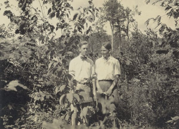 Two boys are standing together against a background of plants and trees. The boy on the right is holding an axe, and the boy on the left is holding some kind of tool. Caption reads: "Henry Thurston - Charles Holt."