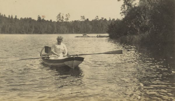 Mrs. Buswell (Mary Emeline "Lina" Porter Buswell) is rowing the boat on Archibald Lake. There are two books behind her on the boat bench. Caption reads: "Mrs. Buswell cheerfully hopes to reach the Hermit."