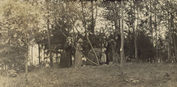 Bending down to earth, Mr. Buswell is planting a small oak tree in honor of his birthday. Mrs. Buswell is on the far left, looking at the camera. Alfred Holt is on the far right, also looking at the camera. Several of the girls are wearing long coats. Caption reads: "Mr. Buswell Planting His Birthday Oak."
