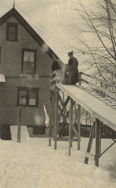 Atop the winter slide, Lillian Wheeler is positioning herself on the sled. Her mother Anna Holt Wheeler is watching her, and a third person is standing behind the sled. There is a house in the background and snow is on the ground. Caption reads: "Mrs. Wheeler and Lillian."