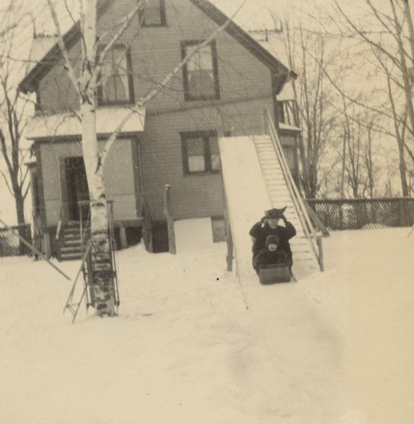 Toddler Lillian Wheeler is tobogganing down the snowy chute with her mother Anna Holt Wheeler, and a third person is sitting behind Anna. There is a house in the background.


