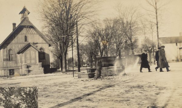 View of the pump and drinking trough for horses on a triangle of land at the junction of Chicago and Main Streets. The first Christian Science Church building in the world is in the background. Three men are walking by the drinking trough. This photograph is one of a series showing the beautification of this small park. Caption reads: "Before: pump and drinking trough for horses."
