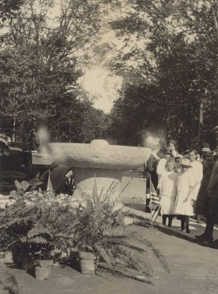View of the new stone drinking fountain for horses at the junction of Chicago and Main Streets. Looking at the camera, two girls are standing to the right of the fountain. There are American flags and potted ferns in the foreground. A car is parked to the left of the fountain. Caption reads: "After: Drinking Fountain Put in by Oconto Women's Club."