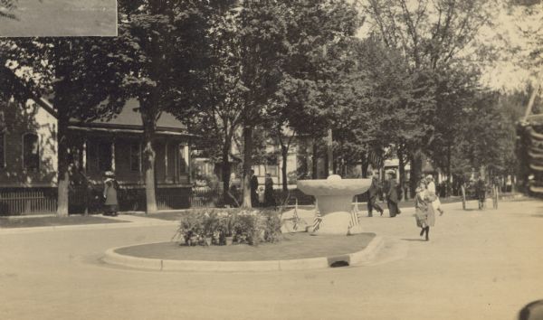 View of the new stone-hewn drinking fountain for horses at the junction of Chicago and Main Streets. A girl is running in the street toward the camera. Small American flags and potted ferns are in the foreground. The road is newly paved. Curb and gutter were recently added. Caption reads: "After: Drinking Fountain Put in by Oconto Women's Club."