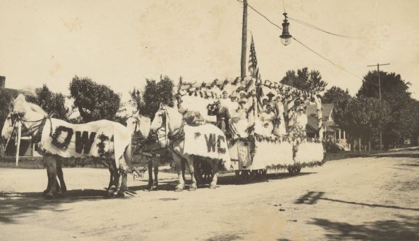 View of a group of mostly women riding on a horse-drawn parade float. The letters "OW" and "WC" are written with flowers on the horse blankets. Several women appear to be nurses. One child is sitting on the float. The driver of the float is tipping his hat.  A man wearing a military uniform is standing on the float. An electric light is hanging over the street. This float was part of the parade celebrating completion of the asphalt paving of Main Street. 


