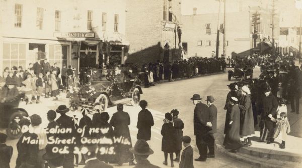 Slightly elevated view of the parade for the dedication of the new Main Street. W.A. Holt is driving his automobile, which is decorated with garlands, in the parade. There are two storefronts on Main Street: the laundry and the ice cream parlor. There is a tall man watching the scene while standing on a tall ladder, which is leaning against a brick building. Men, women and children are lining the sidewalks. Caption reads: "Dedication of new Main Street, Oconto, Wis."