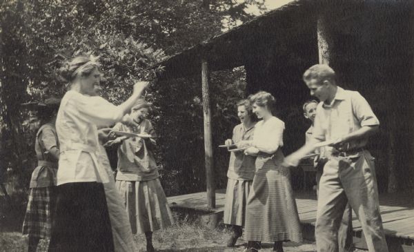 Jeannette Holt, family and friends are laughing and having a watermelon seed fight in front of Island Lodge. Names from left to right: Jeannette Holt, unknown, unknown, Eleanor Holt, Barbara Lyon, Donald Holt, and Phil Smith. Caption reads: "Watermelon Seed Fight. Barbara Lyon. Phil Smith."
