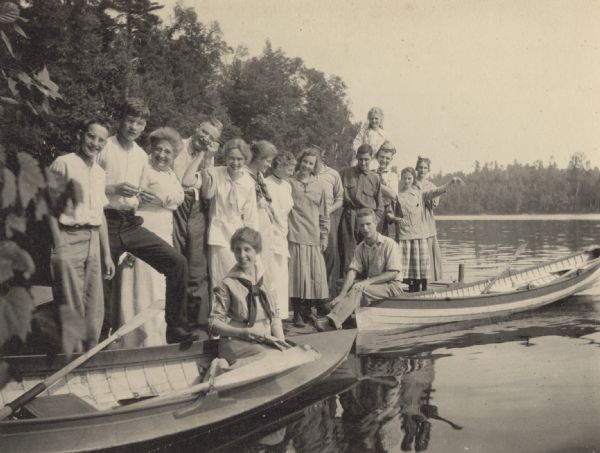 The Holts, Strohs, Buswells, and Wheelers are standing on the Holt pier at Archibald Lake. The Island is in the background. Names from left to right: Donald Holt, Alfred Holt, Mrs. Emeline "Lina" Porter Buswell, Mr. James O. Buswell, Margaret Stroh (?), Harriet Stroh (?), Lucy Rumsey Holt, Eleanor Holt, Oliver Buswell (?), Karl Buswell, Lillian Wheeler (?), Gordon Wheeler (?), Juliet Stroh (?), and Jeannette Holt. In the foreground, Barbara Lyon sitting in a boat, and Phil Smith is sitting on the front of another boat with his feet on the pier. Caption reads: "Holts, Strohs, Buswells, Wheelers, Barbara Lyon, Phil Smith."
