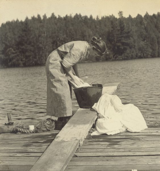 Standing on the pier, Jeannette Holt is doing laundry by hand, using the washboard in an iron bucket that is sitting on the diving board. Archibald Lake and the far shoreline are in the background.