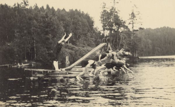 This action shot captures a group on a raft diving into Archibald Lake, with the Island in the background. The divers are probably members of the Holt, Stroh, Buswell, and Wheeler families. There is a slide on the raft.

