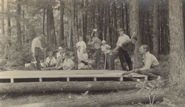 A group of men are building the wood floor for the tented campsite on the Island. In the background, a group of women and children are watching construction. Names of the men from left to right: Unknown, Unknown, Oliver Buswell, Karl Buswell, and Grant Stroh. Names of women and children: Harriet Stroh, Unknown (hat), Barbara Lyon, Margaret Stroh, Eleanor Holt, Jeannette Holt, Anna Wheeler, Lillian Wheeler, unknown (boy with hat). Caption reads: "Building Floor for Tent."