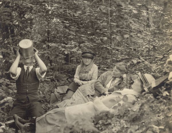 Wearing a hat and scarf, an unknown woman is sitting on the ground in the woods, looking at the camera. To her right, amidst ferns and plants, Lucy Rumsey Holt, wearing a necktie, is lying on her side. Jeannette Holt is lying on her back, looking up. The man on the left is eating or drinking something from a metal pail that is covering his face. There is another pail by Jeannette Holt's feet.
