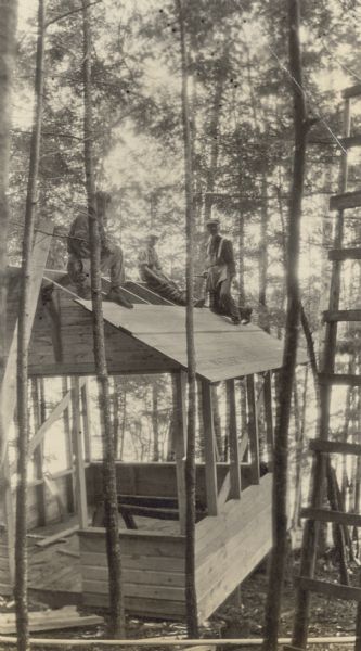 View from hill looking towards three men on the roof while building The Retreat.  There is a tall ladder against a tree on the right. Archibald Lake is in the background. Page heading reads: "Queen's Throne." Caption reads: "Building the Retreat."