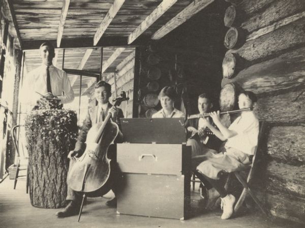 Arch McClure, on the left, is Holding a book and standing behind the home-made wooden podium that is decorated with flowers. A group of musicians is playing for the Sunday church service on the porch at Island Lodge. Names from left to right: Arch McClure, Alfred Holt, Miss Hank, Nathan McClure, and Donald Holt. Caption reads: "Church on Porch at Island Lodge. Arch, Alfred, Miss Hank, Nathan, Donald."

