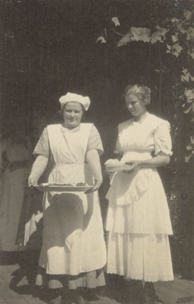 Portrait of two women wearing aprons, possibly cooks. The woman on the left is holding a platter. The woman on the right is holding a stack of small plates. 