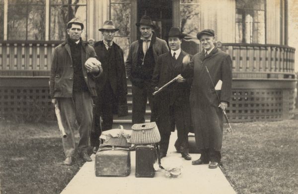 A group of men are standing in front of the Holt family home on Main Street. Several of the men are holding fishing poles, and they appear ready to go on a fishing trip. A pile of luggage, including a wicker fishing creel, is in the foreground. Names from left to right: Rev. John Robertson Macartney, W.A. Holt, Mr. Norman Sleezer, Rev. Ernest W. Wright, W.M. Comstock. Caption reads: "Mr. Macartney, W.A.H, Mr. Sleezer, Rev. Wright, Mr. Comstock."