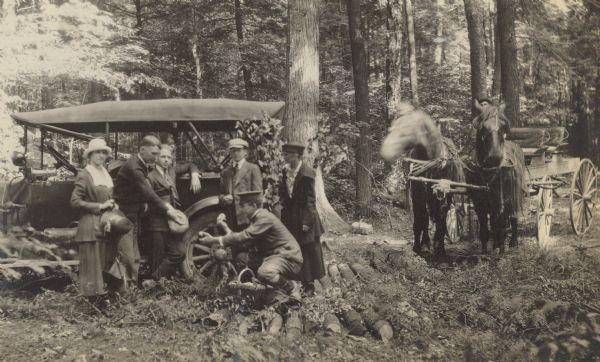 It appears that a set of logs was just placed on the dirt road between an automobile and a horse-drawn carriage, possibly to fix a rut. While squatting by the wicker basket, Phil Smith is handing an apple to W.A. Holt. Standing by the automobile, from left to right: Eleanor Holt, W.A. Holt, Donald DeWitt, Donald Holt, and Jeannette Holt. On the right there is a man sitting in the carriage supervising the horses.
