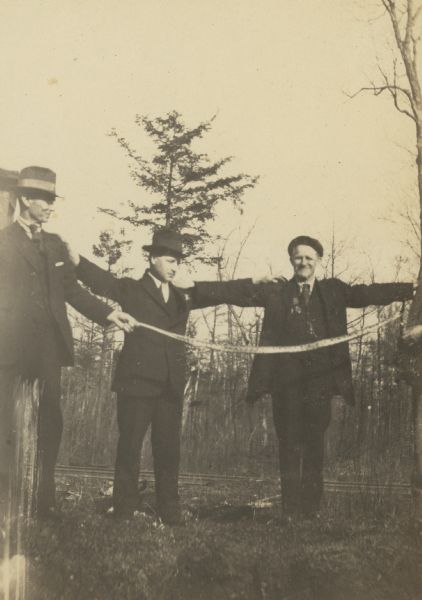 Three men are displaying a very long snake. Names from left to right: W.A. Holt, Rev. Ernest Wright, and W.M. Comstock. Caption reads: "Snake!"