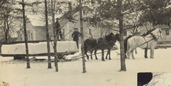 Four horses are pulling the flat bed wagon loaded with ice bricks. One of the men is holding the reins to the horses, and the other man is standing with his hands on his hips. The house in the background is probably the W.A. and Lucy Holt home at 523 Main Street.