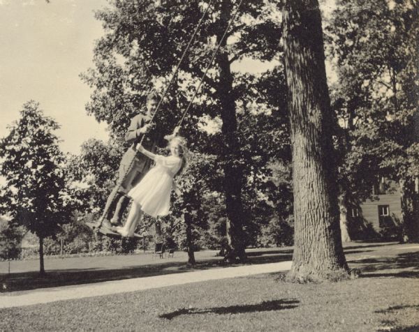 During Word War I, a group of soldiers training at Fort Sheridan visited the home of W.A. Holt's parents. The Holt family entertained the soldiers. Lillian Wheeler is showing the tree swing to one of the soldiers. Many trees and a building are in the background.