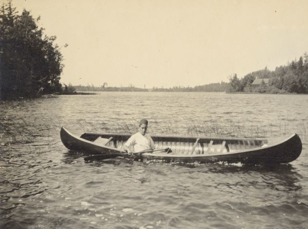 Robert Thurston is canoeing on Archibald Lake amidst some shoreline grasses. The Ark cottage on the Island is in the background on the right.