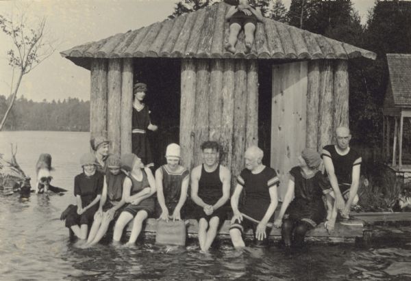Sitting in front of the bath house, bathers are dangling their feet in Archibald Lake. A young man is sunbathing on the roof. A woman is standing in the bath house doorway. A collie, possibly Queenie, is walking in the lake in the background on the left. Names, sitting from left to right: Unknown, Jeannette Holt, Unknown, Unknown, Oliver Buswell (?), I.P. Rumsey, Juliet Rumsey Stroh (?), and Grant Stroh. On the roof: Unknown. In the doorway: Unknown. Page Headline reads: "Aug 13-20."