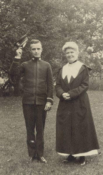 Wearing his military uniform, Phil Smith is doffing his hat to the photographer. Mrs. Smith, with hands interlaced, is standing next to him. A large tree is in the background. Caption reads: "Phil Smith and Mrs. Smith." 
