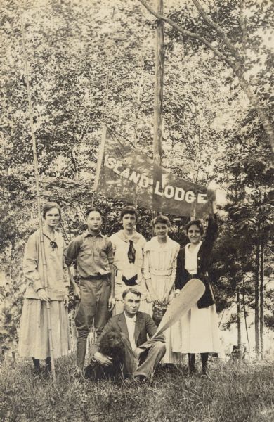 Most of the Lucy and W.A. Holt family are gathered around the Islander flag, which appears to be new. All of the children are present except Alfred Holt. Trees and Archibald Lake are in the background. Standing from left to right: Jeannette Holt, who is holding a fishing pole, Donald Holt, Eleanor Holt, Lucy Rumsey Holt, and Unknown. Sitting: W.A. Holt, who is holding a paddle, and the family dog, a Cocker Spaniel.