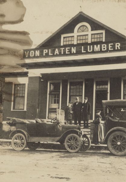 Three men are standing together on the front steps of the Von Platen Lumber building. W.A. Holt is on the right. Presumably one of these men is business owner Godfrey Von Platen. A woman is standing on the sidewalk between two automobiles. There is a dirt road in the foreground.