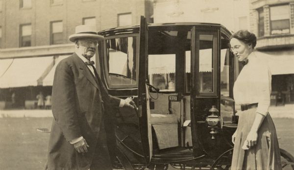 Minnie May Rumsey is standing beside her new Waverley electric car made by the Woods Motor Vehicle Company. Her father Captain I.P. Rumsey, a Civil War veteran, is standing near her and opening the car door. This car has plate glass windows on the front, back, and sides, as well as the door. Caption reads: "Minnie May's new 'Woods'."