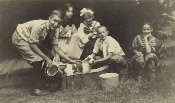 The children are giving a bath to the Holt family dog named Spotty who is standing in a wash tub. Philip Smith is laying his hand on Spotty's back. Alfred Holt is helping Spotty look at the camera. Names from left to right: Philip Smith, Unknown, Jeannette Holt, Alfred Holt, Eleanor Holt.