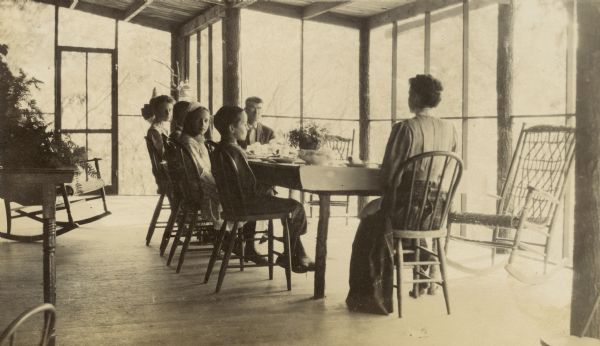 A view of a Holt family lunch or dinner on the porch at Island Lodge. W.A. Holt is sitting at the far end of the table, and Lucy Rumsey Holt is sitting at the near end. Names of the children from left to right: Jeannette Holt, Alfred Holt, Eleanor Holt, and Donald Holt. The children are looking toward Archibald Lake.