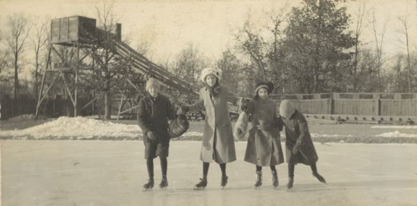 The Holt children are skating in a line at an ice rink. The girls are holding fur hand warmers. There is a toboggan slide in the background. Names from left to right: Alfred Holt, Jeannette Holt, Eleanor Holt, and Donald Holt. Caption reads: "Skating at the Winter Club."