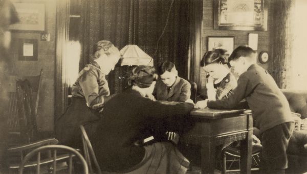 Group of people playing the card game called Flinch. There is an electric lamp in the background. Caption reads: "Cousin Carolyn, foster mother during absence of father and mother between Jan 6th - XXX. Playing Flinch." Names from left to right: Cousin Carolyn, Jeannette Holt, Alfred Holt, Eleanor Holt, and Donald Holt.