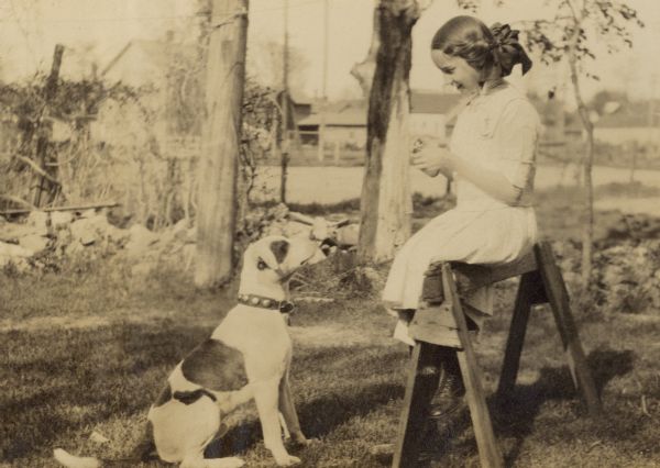 Sitting on a wooden sawhorse in the backyard, Eleanor Holt is giving a treat to Spotty, the family dog.