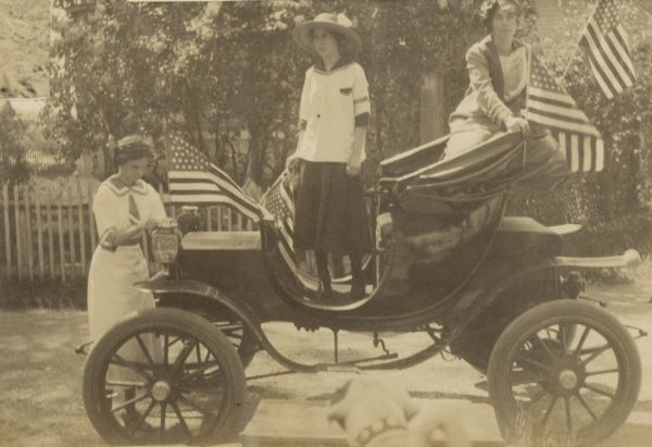 Three girls are decorating Lucy Rumsey Holt's electric car with American flags for Decoration Day, possibly for a parade. Jeannette Holt is on the left. Spotty, the Holt family dog, is in the foreground.