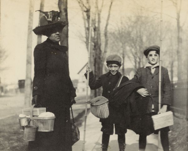Donald Holt and Fred Mooney are going fishing. Each boy is carrying a long bamboo fishing pole. Fred Mooney has the creel. Mrs. Mahoney is carrying a wooden basket with a small bucket and tin cup. Caption reads: "Mrs. Mooney, Fred Mooney, Donald Holt."
