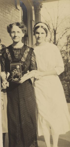 Amateur photographer Lucy Rumsey Holt is holding a folding camera. Her daughter Jeannette Holt is standing beside her outside on the porch at the Evergreens.