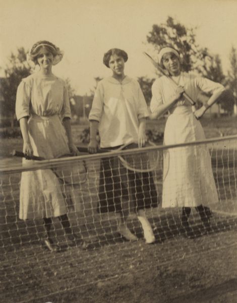 Three tennis players, holding their wooden racquets, are posing at the net on a grass court. Names from left to right: Jeannette Holt, Jean Fergeson, and Ethel Cecil. Caption reads: "Jeannette, Jean, and Ethel."