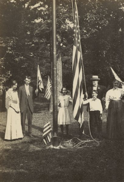 At the home of Civil War Captain Israel Parsons Rumsey, the group is raising the American flag on Independence Day. Names from left to right: Lucy Rumsey Holt, W.A. Holt, Eleanor Holt, Captain I.P. Rumsey, Donald Rumsey Holt, and Mary Matilda Rumsey. Caption reads: "Raising the Flag at Grandfather's."