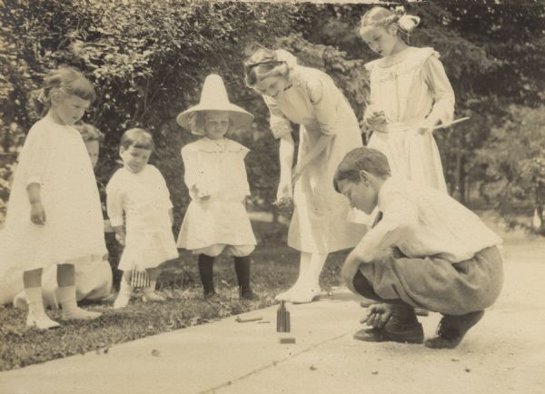 Alfred Holt is setting off some firecrackers while his sisters and several cousins are watching. Names from left to right: Elizabeth Doud Rumsey, Marion Edgerton Doud Rumsey, Henry Axtell Rumsey, Jr., Lillian Wheeler, Jeannette Holt, Alfred Holt, and Eleanor Holt. Caption reads: "Elizabeth, Henry, Lillian, Jeannette, Alfred, Eleanor."
