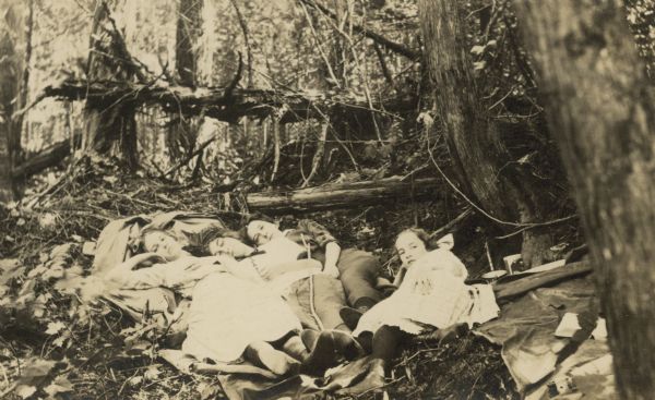 Jeannette Holt and two close friends are lying down on their backs on a tarp next to each other in the woods. Younger sister Eleanor Holt is also lying down, next to Jean Fergeson. A fallen tree and various plants are in the background. An open book, a tin cup, a canister, and possibly a camera are on the right. Names from left to right: Jeannette Holt, Ethel Cecil, Jean Fergeson, and Eleanor Holt. Caption reads: "Jeannette, Ethel Cecil, Jean Fergeson."