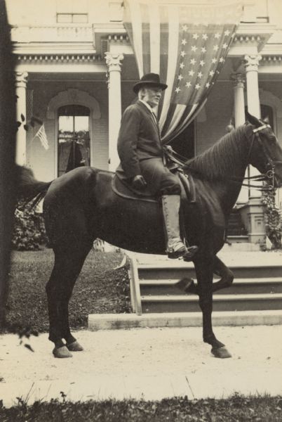 Civil War veteran Captain Israel Parsons Rumsey, the father of the photographer, is sitting on a horse in front of his home. A large American flag, hanging from the second floor balcony, is behind Capt. Rumsey. The caption reads: "Grandfather Rumsey - Fourth of July."