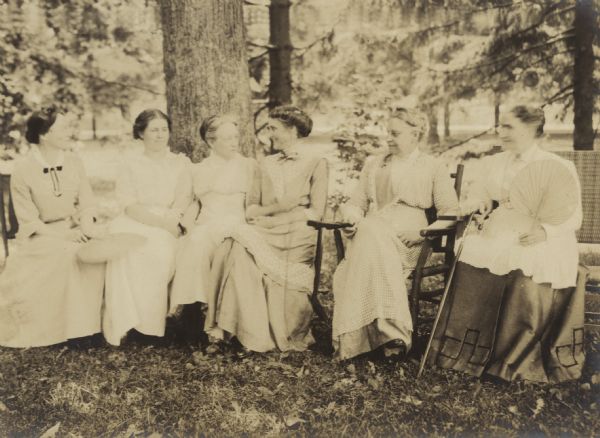 Two generations of women sitting together and socializing under a big tree. Two women are holding hand fans. The theme of this image seems to be sisterhood. Names from left to right: Mary Mathilda Rumsey, Edna Rumsey, Caroline {unknown}, Juliet Rumsey Stroh, Dena Turnley, Mary Mathilda Axtell Rumsey. Caption reads: "M.M. Rumsey, Edna Rumsey, Cousin Caroline, Juliet Rumsey Stroh, Aunt Dena Turnley, Grandmother Rumsey."