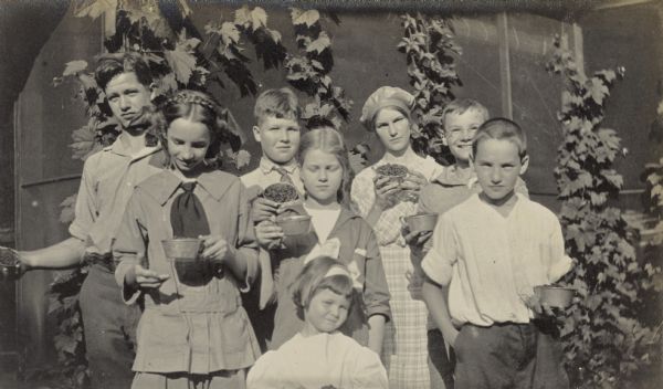 After berry picking, the group of Trowbridges and Holts display their tin cups filled with local fruits. Names from left to right: Calvin Trowbridge, Eleanor Holt, Corneil Trowbridge, Katherine Trowbridge, Jane Trowbridge, Jeannette Holt, Alfred Holt, Donald Holt. Caption reads: "Trowbridges and Holts. Calvin, Corneil, Katherine, Jane."