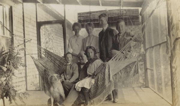 The family is posing on the screen porch at Island Lodge. Eleanor Holt, wearing a leg brace, is sitting on the fringed hammock with her maternal grandfather Captain Israel Parsons Rumsey, and her younger brother Donald Holt. Family dog Spot is sitting on the floor next to Donald Holt. Standing behind the hammock (from left to right) is Jeannette Holt, Lucy Rumsey Holt, W.A. Holt, and Alfred Holt.
