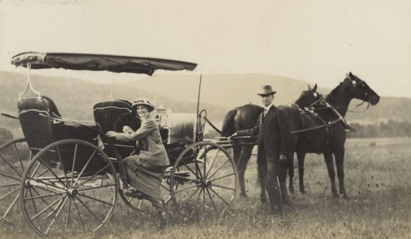 Jeannette Holt is smiling and stepping into the horse-drawn carriage with a top. Her father W.A. Holt is holding the horses' reins. There are hills in the background.