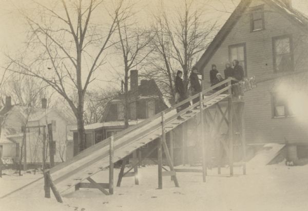 A group of children and adults is on the wooden toboggan slide in the backyard of the Lucy and W.A. Holt home. One person on the top platform is holding a toboggan. Several others are also waiting up top. Two people are climbing up the stairs beside the slide. Caption reads: "Toboggan Slide in Backyard."
