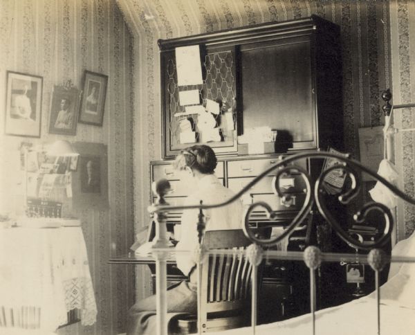 With her side to the camera, Lucy Rumsey Holt is sitting at her desk in her bedroom. Sunlight is streaming in from the window on the left. The bedroom walls are covered with floral wallpaper. A book and a lamp are on top of the table in front of the window. Framed photographs, including ones of her father and her husband, are hanging on the wall. Additional photographs and a calendar are pinned on the cork board. The foot of the metal 4-post bed is in the foreground. The Oconto directory is hanging on the side of the desk. Caption reads: "Lucy Holt at her desk in her bedroom."